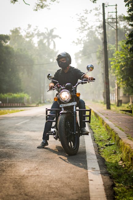 The image shows a person wearing a black helmet, face mask, and casual dark clothing riding a black motorcycle along a quiet outdoor street during daytime. The motorcycle is positioned on the right si