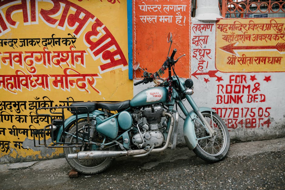 A vintage light blue Royal Enfield motorcycle parked against a colorful wall featuring yellow, red, and blue signs with Hindi and English text. The motorcycle has a black seat, chrome exhaust pipe, an