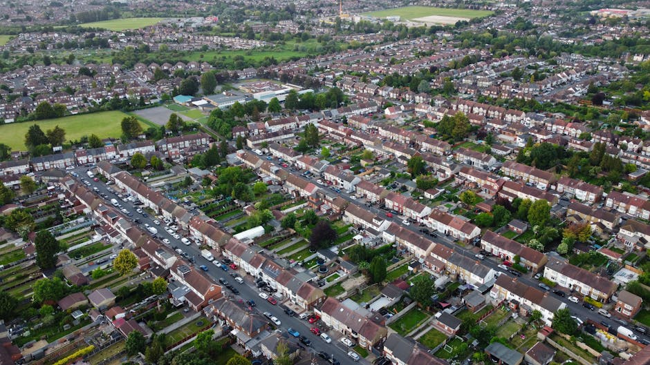 An aerial view of a residential neighborhood showing a grid of terraced houses with tiled roofs, small gardens and backyards, and tree-lined streets with parked cars. The area features well-maintained