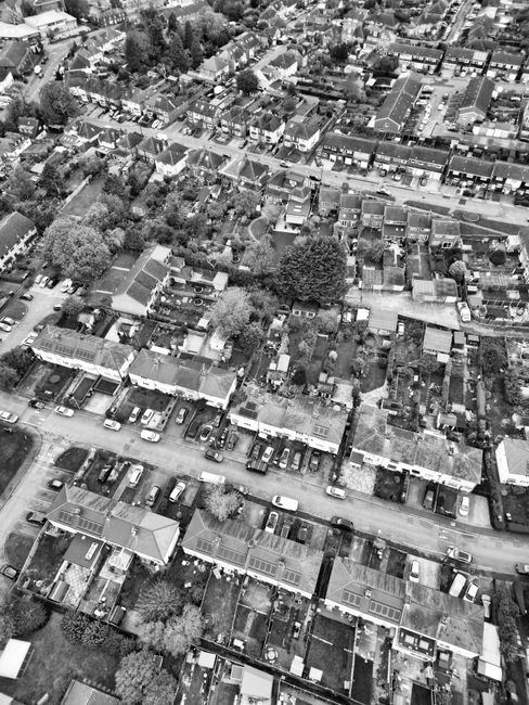 An aerial view of a residential neighborhood showing a grid of terraced houses with tiled roofs, small gardens and backyards, and tree-lined streets with parked cars. The area features well-maintained